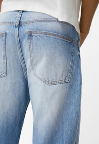 Ljusblå denimjeans med synliga bakfickor, bärs av en person i vit skjorta, visad från midjan neråt bakifrån.