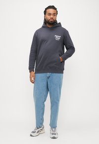 Σκούρο γκρι φούτερ με κουκούλα και τσέπη μπροστά, με λευκές λεπτομέρειες λογότυπου, σε συνδυασμό με ανοιχτό μπλε φαρδιά τζιν και γκρι αθλητικά παπούτσια. Ουδέτερο φόντο.