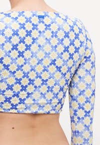 Long-sleeve cropped top featuring a blue fabric with a floral pattern in yellow and green. Tag displayed at the back. Smooth texture.
