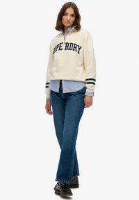 Crème sweatshirt met "Superdry" in marineblauw, ritssluiting en zwarte strepen op de mouwen. Gelaagd over een blauw-roze gestreept shirt en blauwe jeans.
