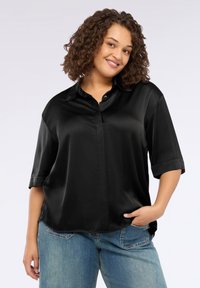 Donna sorridente con capelli ricci, indossa una blusa nera in raso larga e jeans blu, mentre posa con una mano in tasca su uno sfondo bianco.