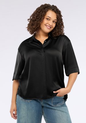 Femme souriante aux cheveux bouclés portant un blouse noire en satin ample et un jean bleu, posant avec une main dans la poche sur fond blanc.
