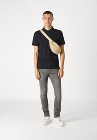 Black polo shirt, light beige sling bag, gray slim-fit jeans, white sneakers, standing against a plain white background.