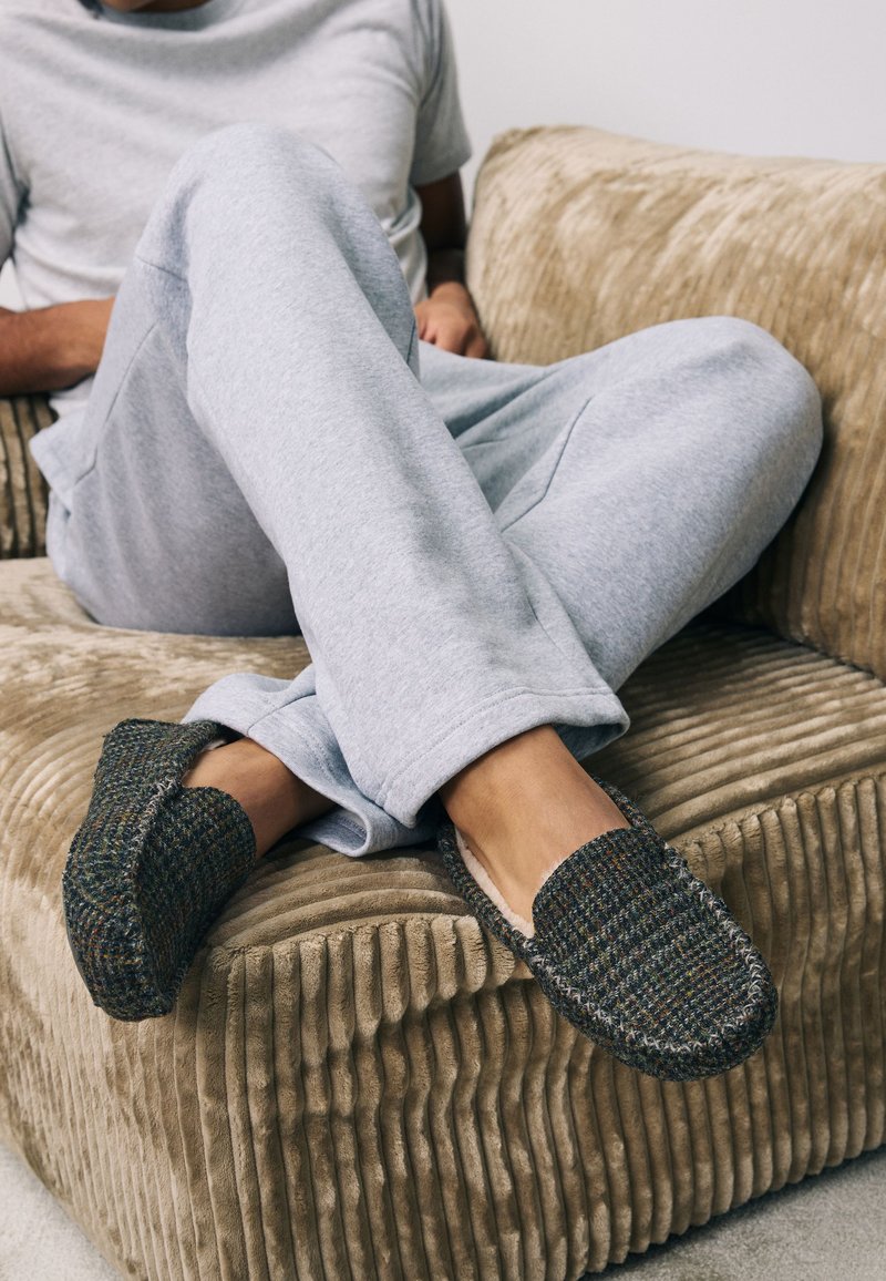 Personne assise sur un canapé en velours côtelé beige portant un pantalon de survêtement gris et des chaussons tissés foncés à enfiler avec des coutures blanches.