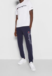 Bílé bavlněné tričko s černým logem "Champion", doplněné tmavě modrými tepláky s vyšitým logem. Celý outfit doplňují bílé sportovní boty.