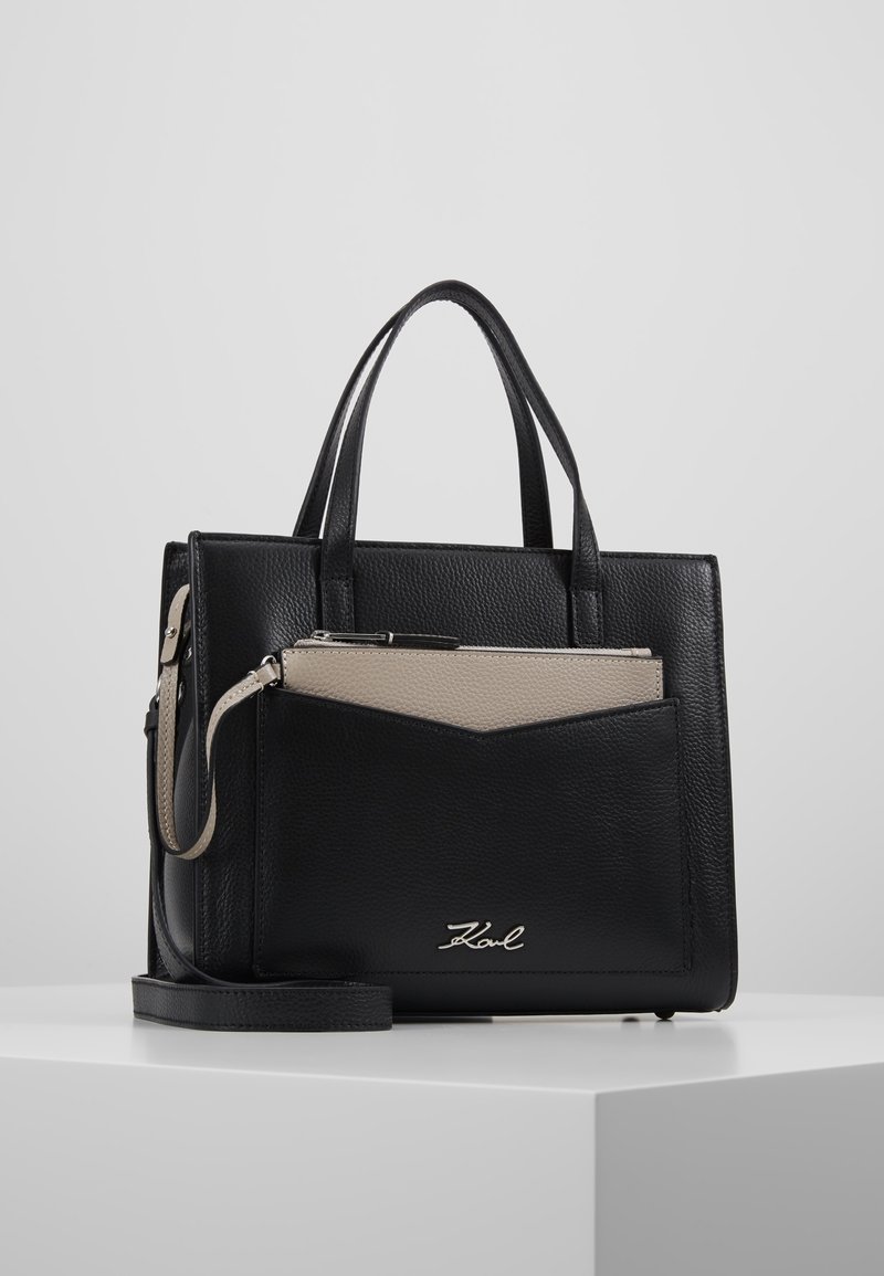 Sac à main en cuir noir avec deux poignées, poche avant contenant une pochette beige zippée et un logo "Karl" argenté à l'avant.