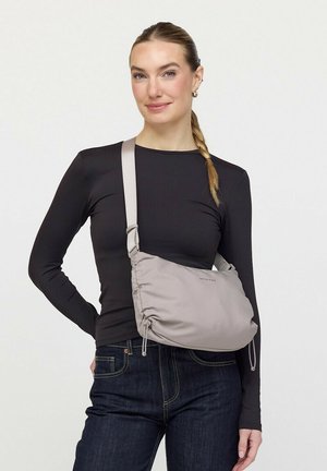 Vrouw met blond gevlochten haar, gekleed in een zwart shirt met lange mouwen en donkere spijkerbroek, draagt een lichtgrijze crossbodytas met verstelbare riem.
