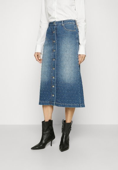 Denim-Midirock mit Knopfleiste und silbernen Nietenakzenten, in einem verblassten Blauton und mit geradem Saum. Kombiniert mit schwarzen Stiefeletten.