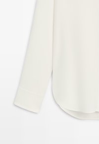 Bílá halenka s hladkou texturou, s dlouhými rukávy a zaobleným lemem. Manžeta má jednoduchý skládací design.