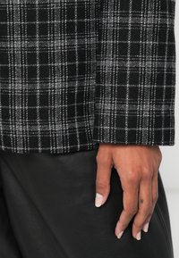 Crno-siva karirana tkanina s mekom teksturom, koja ima kroj uz tijelo i ravne rukave. Ruka se oslanja na kožnu površinu.