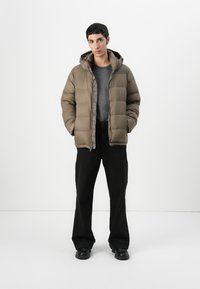 Puffer jacket in olive green, featuring a hood, quilted design, and front zip closure, paired with black trousers and shoes.