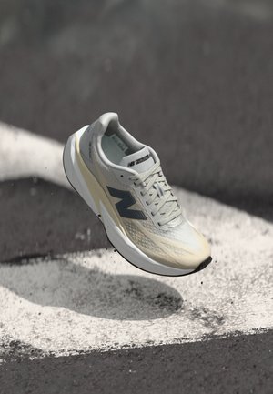 Lys grå og beige New Balance joggesko i luften over asfalt med hvite veimerker.