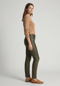 Vrouw staand in een beige trui, olijfgroene leren broek en bruine loafers tegen een effen lichtgrijze achtergrond.