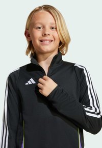 Svart träningsjacka med vita ränder, hög krage och figursytt snitt; har Adidas-logotyp på bröstet och kontrasterande lila accenter.