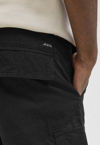Black cargo pants with a matte texture, featuring a patch pocket and a logo label. Designed with a relaxed fit and practical pockets.