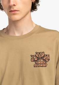Beige cotton t-shirt featuring a graphic of three bear heads and the text "TIMBER ELEMENT" in bold black and orange below the illustration.