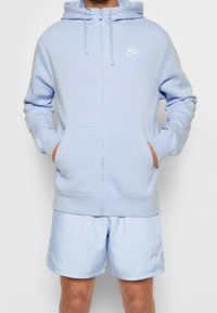 Ljusblå zip-up hoodie i mjukt tyg, med en framficka, dragsko och en liten vit Nike-logotyp på bröstet.