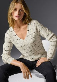 Blonde Frau, die auf einem weißen Stuhl sitzt, trägt einen cremefarbenen, gemusterten V-Ausschnitt-Pullover, schwarze Hosen und ein silbernes Armband vor einem grauen Hintergrund.