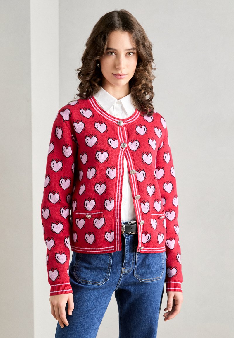 Punainen neulevillatakki, jossa on vaaleanpunaisia sydäntulostuksia, napitettavalla etuosalla, pyöreällä kaula-aukolla ja kontrastivalkoisilla reunuksilla. Kaksi etup distinctiä taskua.