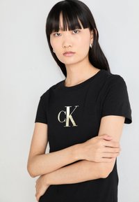 Melna kokvilnas T-krekla ar īsām piedurknēm priekšā, kurā ir metāliskas zeltītās "cK" logo. Audums izskatās gluds un pieguļošs.