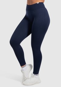 Marinblå leggings med hög midja, tillverkade av elastiskt material, med slät textur och en åtsittande passform, tillsammans med vita sneakers.