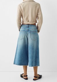 Giacca beige corta, culotte in denim blu, vestibilità rilassata, effetto sbiadito, orlo sfrangiato, tasche posteriori e sandali neri piatti, visti da dietro.