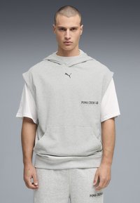 Άνδρας με ξυρισμένο κεφάλι, φοράει αμάνικο φούτερ ελαφρώς γκρι πάνω από λευκό μπλουζάκι και ταιριαστό φόρμα με λογότυπα Puma, στέκεται μπροστά σε γκρι φόντο.