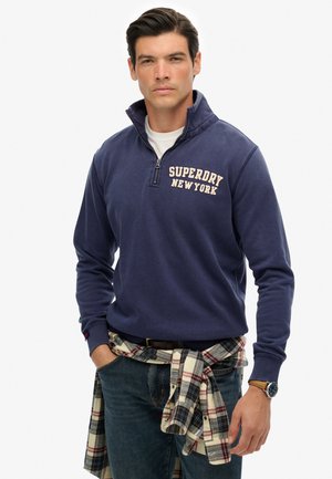 Navy blauwe zip-up pullover met "SUPERDRY NEW YORK" logo in lichtbeige. Katoenmix stof, lange mouwen, gecombineerd met een geruit flanellen overhemd om de taille.