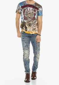 Camiseta gráfica de manga corta para hombre con un diseño colorido que presenta un cráneo de lobo, combinada con unos vaqueros claros y desgastados con costuras detalladas.