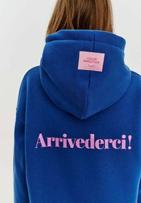 Modrá mikina s kapucí vyrobená z měkké látky, s růžovým grafikou se slovy "Arrivederci!" na zádech, s růžovou nášivkou na výstřihu.