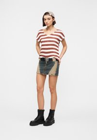 Γυναίκα που φοράει μπεζ καπέλο προς τα πίσω, κόκκινο και άσπρο ριγέ κοντομάνικο πουκάμισο, δίχρωμη μίνι φούστα από τζιν με ζώνη σε σχήμα καρδιάς, και μαύρα μποτάκια μάχης.