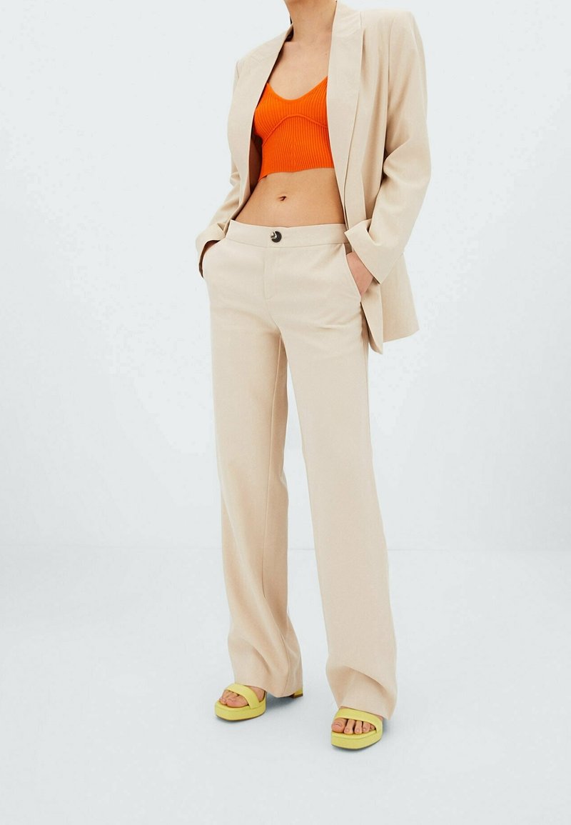 Pantalon beige sur mesure avec une texture lisse, associé à un crop top côtelé orange et des sandales plateforme jaunes, ajoutant un contraste de couleurs.