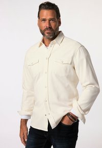 Kermanvärinen pitkähiha-paita, jossa painonapit, kaksi rintataskua ja kevyt tekstuuri. Hihat käärityt ylös, päällä tummansiniset farkut.