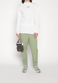 Long-sleeve white shirt with black Nike logo, green pants, teal sneakers, and brown shoulder bag. Casual outfit with sporty elements.