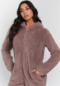 Un mono con capucha marrón, suave y esponjoso, de manga larga y con bolsillos laterales. La tela tiene una textura suave y un aspecto de peluche. Cierre frontal con cremallera.
