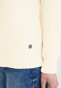 Krämfärgad stickad tröja med en grov textur, med en svart logotypdetalj på sidan, kombinerad med blå denimjeans.