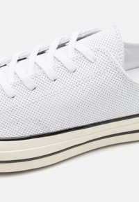 Bílé perforované tenisky s kulatou špičkou, plochými tkaničkami a černým pruhem podél gumové podrážky. Materiál vypadá jako lehký a texturovaný.