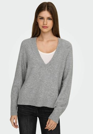 Grå v-ringad tröja gjord av mjukt tyg med avslappnad passform, ribbade muddar och nederkant. Buren över en vit topp, i kombination med mörka jeans.