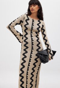 Bež triiks vormiga kleit, millel on lainelised mustad mustrid, pikad varrukad laienevate äärtetega ja lihvitud siluett. koos musta kotiga.