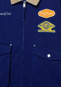 Modrá bunda s béžovým límcem, zipem, dvěma předními kapsami s klopami a třemi nášivkami s nápisy „Pull Records“, „Records California State 1991“ a „Neighborhood Club“.