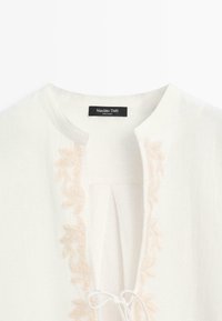 Blouse blanche avec broderie florale beige le long de l'encolure, col rond et petite fermeture à nouer au centre devant.