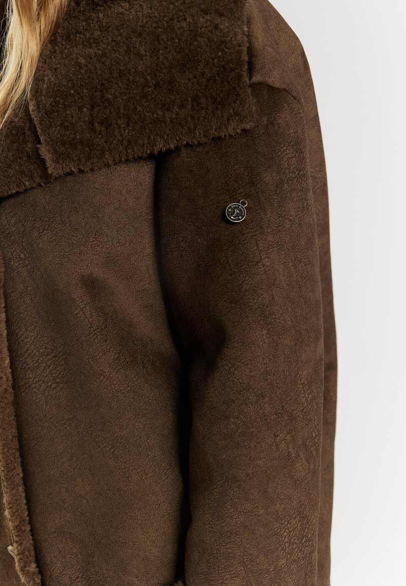 Bruine, getextureerde jas met een zachte bruine bontkraag en een ronde merklabel op de mouw. Het materiaal heeft een suède-achtige uitstraling.