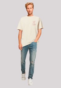 Beige graphic t-shirt with short sleeves and a cartoon print on the chest, paired with distressed light blue jeans and white sneakers.