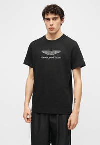 Νέος άνδρας με κοντά σγουρά μαλλιά, φορώντας μαύρο T-shirt της ομάδας Aston Martin Formula One και μαύρο παντελόνι, με απλό φόντο.