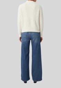 Eine Person trägt einen weißen strukturierten Pullover und lockere blaue Jeans und steht mit dem Rücken zur Kamera vor einem neutralen Hintergrund.