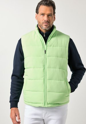 Mand iført lysegrøn quiltet vest med lynlås over sort langærmet skjorte og hvide bukser, stående foran en ensartet lys baggrund.