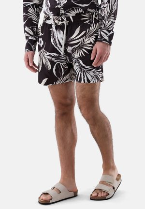 Mann trägt schwarz-weiße Shorts mit tropischem Muster und passendes langärmeliges Oberteil mit beigen Sandalen, steht vor weißem Hintergrund.