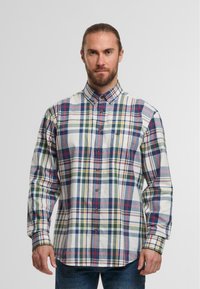 Mann mit Bart und zusammengebundenem Haar, der ein langärmeliges, kariertes Hemd in mehreren Farben und blaue Jeans trägt, vor einem einfarbigen Hintergrund.