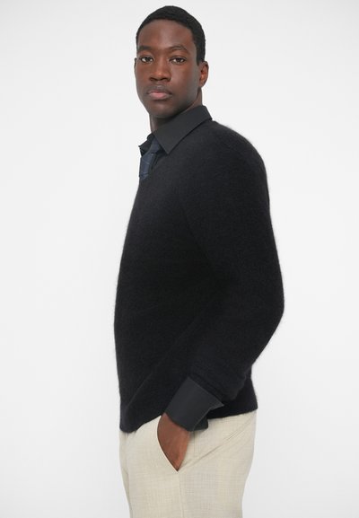 Pullover nero fluffy con scollo a V, indossato sopra una camicia nera con colletto e pantaloni color crema. La texture sembra morbida e voluminosa.