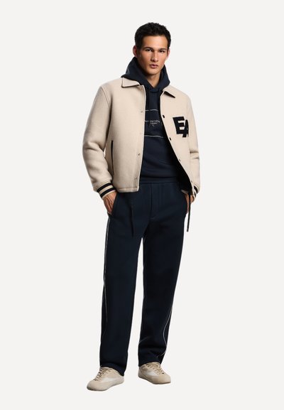 Junger Mann steht mit den Händen in den Taschen, trägt eine beigefarbene Collegejacke, einen navyblauen Hoodie, passende Hosen und beiges Sneakers auf weißem Hintergrund.